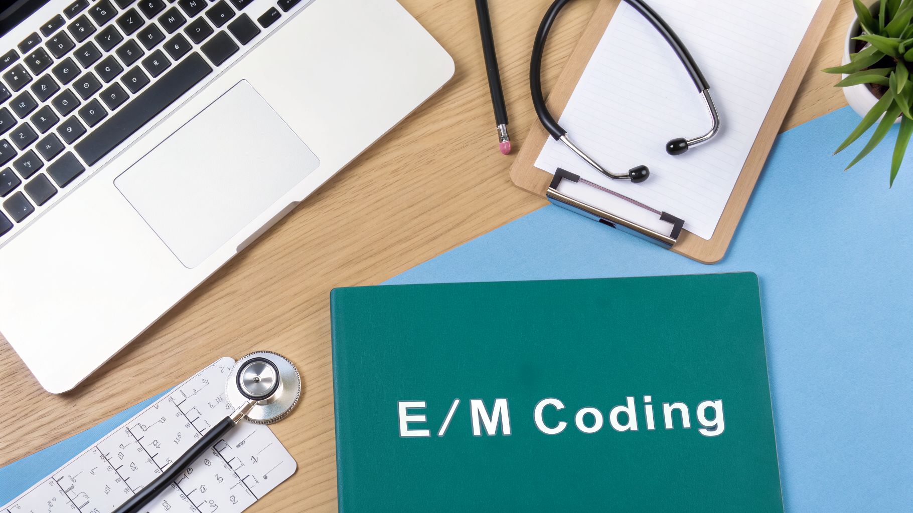 A desk flat lay with a laptop, stethoscope, and a green folder labeled 'E/M Coding'.